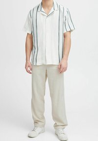 Man in een wit overhemd met korte mouwen en verticale strepen, beige broek, witte sneakers en een zilveren armband om zijn linkerpols.