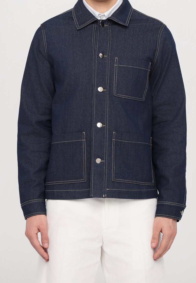 Homme portant une veste en denim bleu foncé avec fermeture à boutons et trois poches plaquées sur le devant, associée à un pantalon blanc.