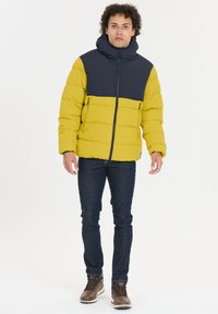 En puffjacka med gul kropp och marinblått ovre, med hög krage, dragkedjefickor och horisontellt quiltat mönster. Bärs med mörka jeans och bruna stövlar.