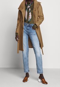 Manteau en laine couleur beige sur un haut à manches longues vert, jeans droits bleu clair, et chaussures marron texturées à enfiler avec un talon blanc. Écharpe à motifs.
