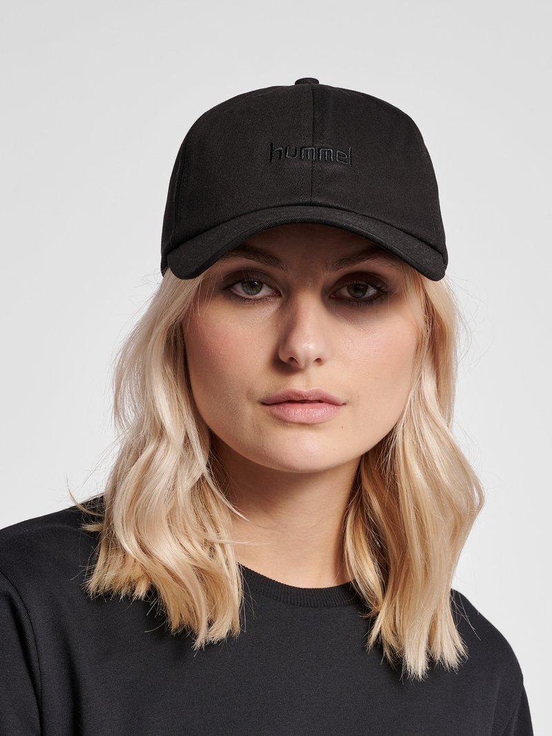 Junge Frau mit blondem Haar trägt eine schwarze Kappe mit "hummel"-Logo und ein schwarzes Shirt und blickt nach vorne vor einem einfarbigen hellen Hintergrund.