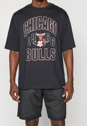 T-shirt noir oversize montrant le logo des Chicago Bulls avec des fleurs et le texte "1966", porté avec un short en mesh noir sur une personne debout.
