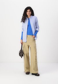 Chemise à rayures bleues et blanches boutonnée sur un haut à manches longues bleu, accompagnée de pantalons cargo larges couleur kaki et de bottines noires.