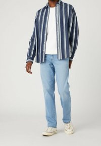 Man som bär ljusblå jeans, vita sneakers, vit t-shirt och en marinblå knäppt skjorta med vita vertikala ränder.