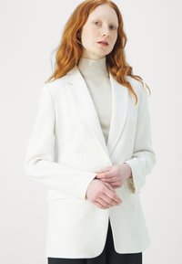 Blazer bianco dal taglio sartoriale, con rever a punta, chiusura con un bottone e tasche laterali, abbinato a un dolcevita beige chiaro.