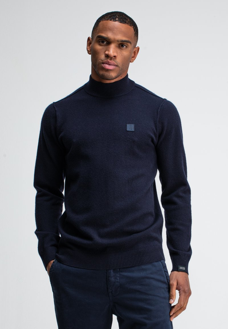 Navyblauer Rollkragenpullover aus einem weichen, strukturierten Stoff mit tailliertem Schnitt sowie gerippten Bündchen und Saum, ergänzt durch ein kleines Logo-Patch auf der Brust.