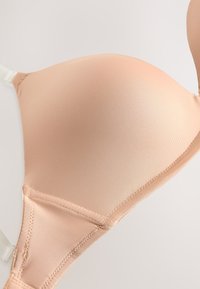 Hladká, svetlo béžová vystužená podprsenková košíčka s priehľadnými nastaviteľnými ramienkami a podšitými švami na neutrálnej farbe pozadia.