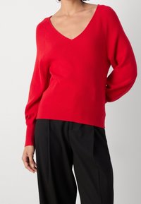 Czerwony sweter w serek z długimi, zwężającymi się rękawami i prążkowaną fakturą. Nosi się go z luźnymi czarnymi spodniami, co podkreśla swobodny krój.