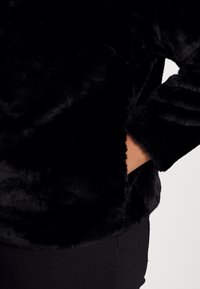 Zwarte pullover van imitatiesbont met een zachte, gestructureerde oppervlakte, lange mouwen en een zak aan de voorkant, gecombineerd met smalle zwarte broek.