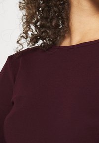 Bordeauxfarbener Stoff-Oberteil mit glatter Textur und Rundhalsausschnitt. Das Material scheint dehnbar zu sein und zeigt subtile Kurven an der Schulter und dem Kragen.