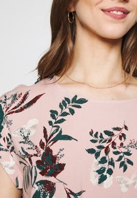 Floral-patterned pink blouse with short sleeves. Features green and burgundy leaves and butterflies. Delicate gold necklace visible.