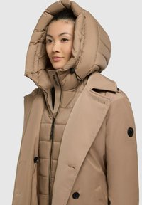 Cappotto esterno beige con un ampio cappuccio imbottito e chiusura con zip, sovrapposto a una giacca interna trapuntata con design leggero e dettagli con bottoni.