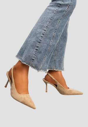 Personne portant un jean court bleu clair et des escarpins slingback à bout pointu en daim beige sur un fond uni.