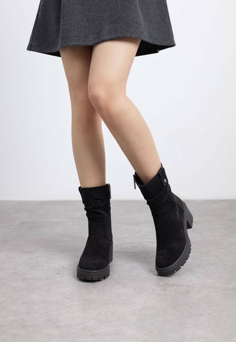 Botines de ante negros con un ligero tacón, que presentan un cierre lateral y suela texturizada para agarre, mostrados con una falda gris en un entorno neutral.