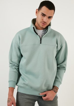 Sudadera de cuello redondo en un suave color verde menta, con media cremallera, puños y dobladillo acanalados. Textura lisa, corte relajado y una única costura horizontal en el pecho.