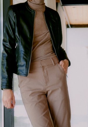 Person wearing a black leather jacket, beige turtleneck, and beige pants, standing indoors with one hand in pocket.