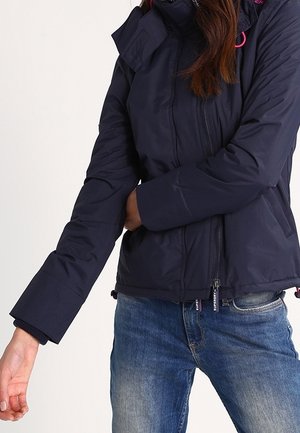 Mujer con chaqueta de invierno azul marino con cremallera frontal y vaqueros azules, ajustando la manga con la mano izquierda.