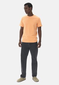 Orange short-sleeve t-shirt, relaxed fit, paired with dark gray jeans. Model wears beige slip-on shoes, standing against a plain background.