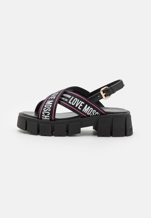 Sort platåsandal med tyk sål med mønster, krydsede stropper med teksten "Love Moschino" og en guldbøjle ankelrem.