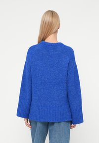 Μπλε πλεκτή μπλούζα με χαλαρή εφαρμογή, ριγωτό τελείωμα και φαρδιές μανσέτες. Η υφή είναι απαλή και χνουδωτή, που προσθέτει στην άνετη εμφάνισή της.