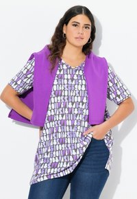Paarse mouwloze cardigan over een kort mouwtjes grijs topje met een geometrisch patroon in wit en paars. Donkere jeans maken de outfit compleet.