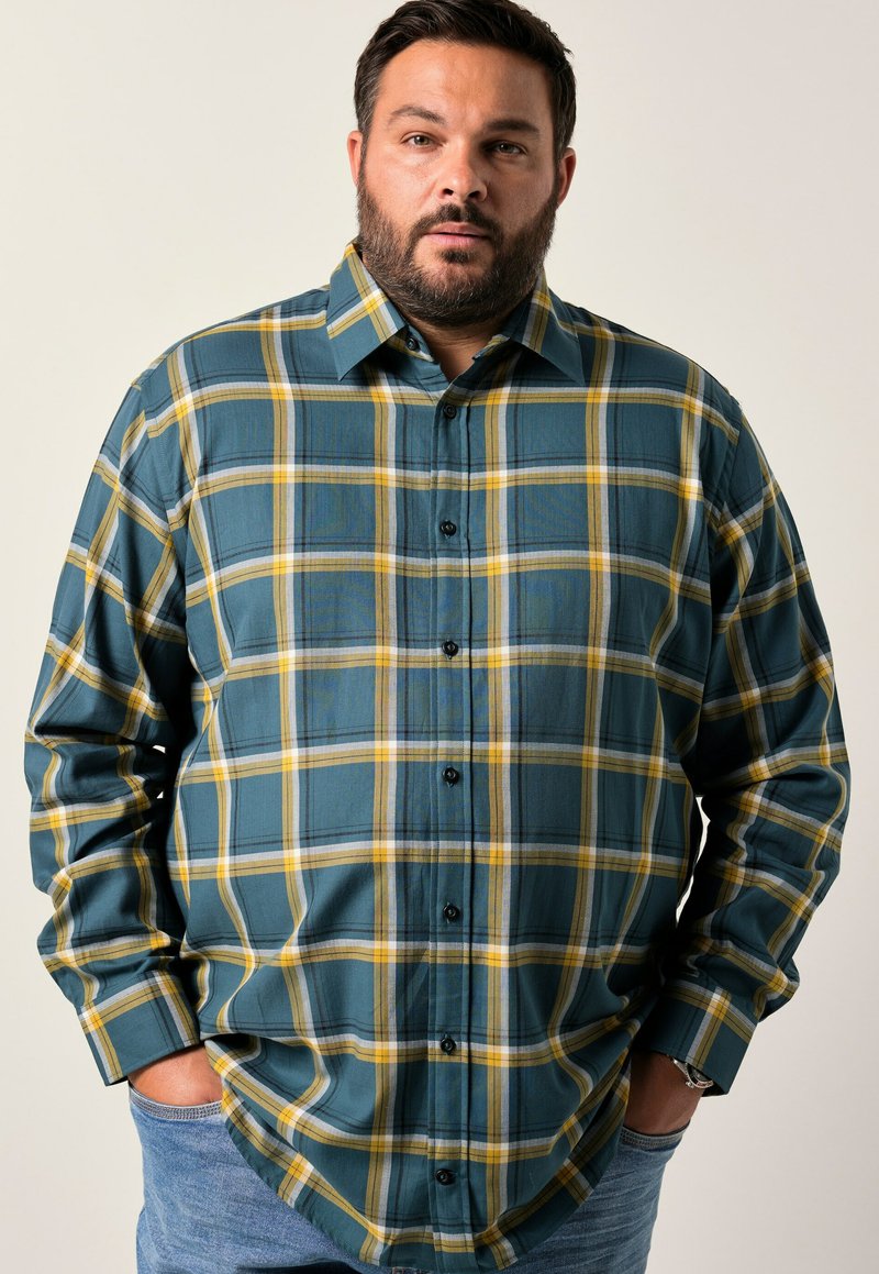 Camicia a quadri verde e gialla con maniche lunghe e bottoni neri, realizzata in un tessuto testurizzato. Indossata su jeans blu.