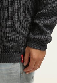 Grijze gebreide trui met geribbelde textuur. Bevat een zwart label en een rood accentlabel aan de zijkant. Mouwdetail zichtbaar op de hand.