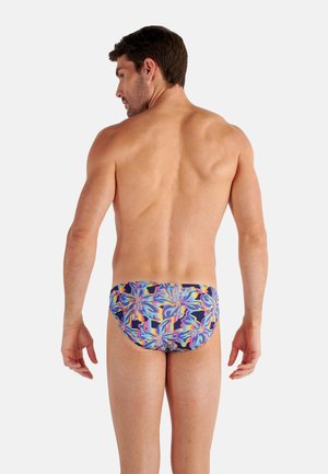 Man standing facing away, wearing colorful swim briefs with abstract floral and geometric patterns on a white background.