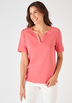 Femme aux cheveux foncés portant un haut corail rose à manches courtes avec des détails à motifs sur les épaules et un pantalon blanc, souriante et regardant sur le côté.