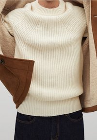 Kremfarvet strikket sweater med ribbet tekstur og rund hals, båret under en brun ruskindsjakke med pelsfoer og knapdetalje.