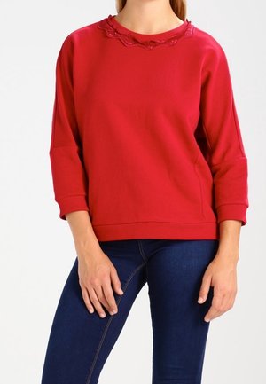 Frau trägt ein rotes Sweatshirt mit langen Ärmeln und Spitzenbesatz am Ausschnitt sowie dunkelblaue eng anliegende Jeans, steht entspannt mit den Händen.