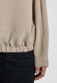 Maglia beige a maniche lunghe con orlo elasticizzato arricciato, indossata da una persona con pantaloni scuri, con la mano rilassata lungo il fianco su uno sfondo neutro.