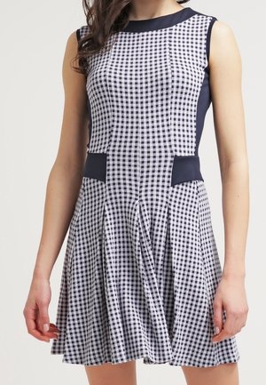 Sleeveless navy and white checkered dress with fitted bodice, navy side panels, and flared pleated skirt worn by a standing person.