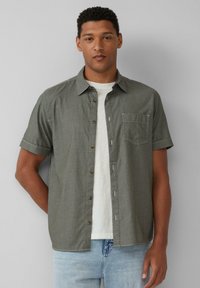Korte mouw knopshirt in olijfgroen textiel, met een borstzak en bruine knopen, gedragen over een wit T-shirt.