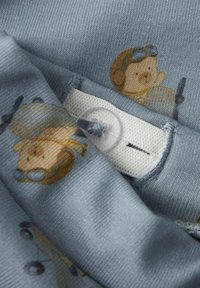 Hellblauer Stoff mit strukturiertem Oberflächendesign, der ein sich wiederholendes Muster von cartoonhaften Charakteren zeigt, dazu ein weißes Etikett und ein transparenter Knopf.
