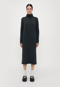 Kullsvart turtleneck-kjole, strikket tekstur, lange ermer, ankellang, med sideslisser, parret med hvite sokker og kraftige svarte sko.