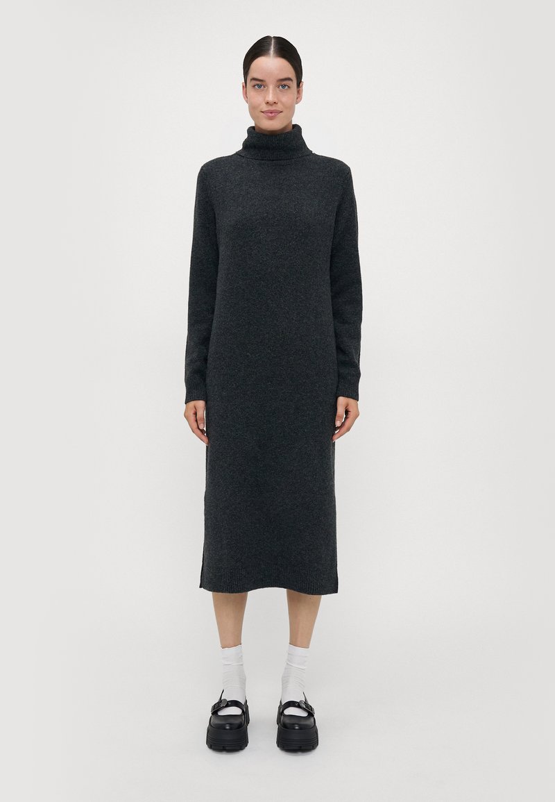 Kullsvart turtleneck-kjole, strikket tekstur, lange ermer, ankellang, med sideslisser, parret med hvite sokker og kraftige svarte sko.
