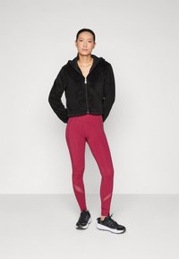 Giacca nera con zip in pelliccia sintetica abbinata a leggings bordeaux con dettagli in rete e scarpe sportive nere con suole bianche.