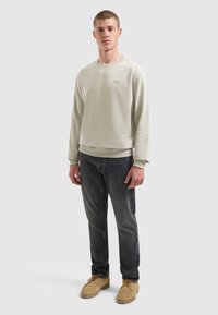 Junger Mann, der einen hellbeigen Pullover, dunkle graue Jeans und tanfarbene Wildlederstiefel trägt, vor einem schlichten hellen Hintergrund.