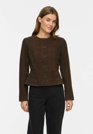 Femme portant une veste marron foncé cintrée boutonnée avec un bas peplum et un pantalon noir, debout devant un fond blanc uni.