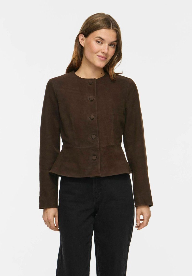 Femme portant une veste marron foncé cintrée boutonnée avec un bas peplum et un pantalon noir, debout devant un fond blanc uni.