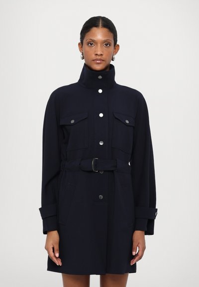 Manteau ceinturé bleu marine avec un col haut, deux poches poitrine à rabat, des poches latérales, et des boutons en argent pour la fermeture et les accents.