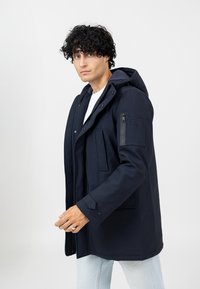 Marineblaue wasserdichte Parka mit Kapuze, vorderer Knopfleiste, mehreren Taschen und Reißverschlussakzent am Ärmel. Leichtgewichtig mit glatter Textur.