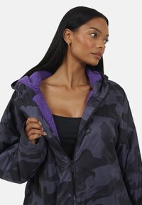 Donna con lunghi capelli scuri che indossa una giacca mimetica nera e viola scuro con un interno in pile viola, parzialmente aperta, che guarda verso destra.