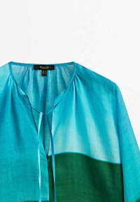 Chemise dégradée turquoise et verte avec col en V, fermeture à nouer, et épaules froncées. Tissu léger et transparent avec une texture lisse.