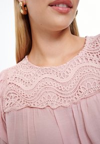 Blusa rosa con dettagli di pizzo intricati nel colletto, texture in tessuto morbido e maniche arricciate. Orecchini a cerchio dorati visibili.