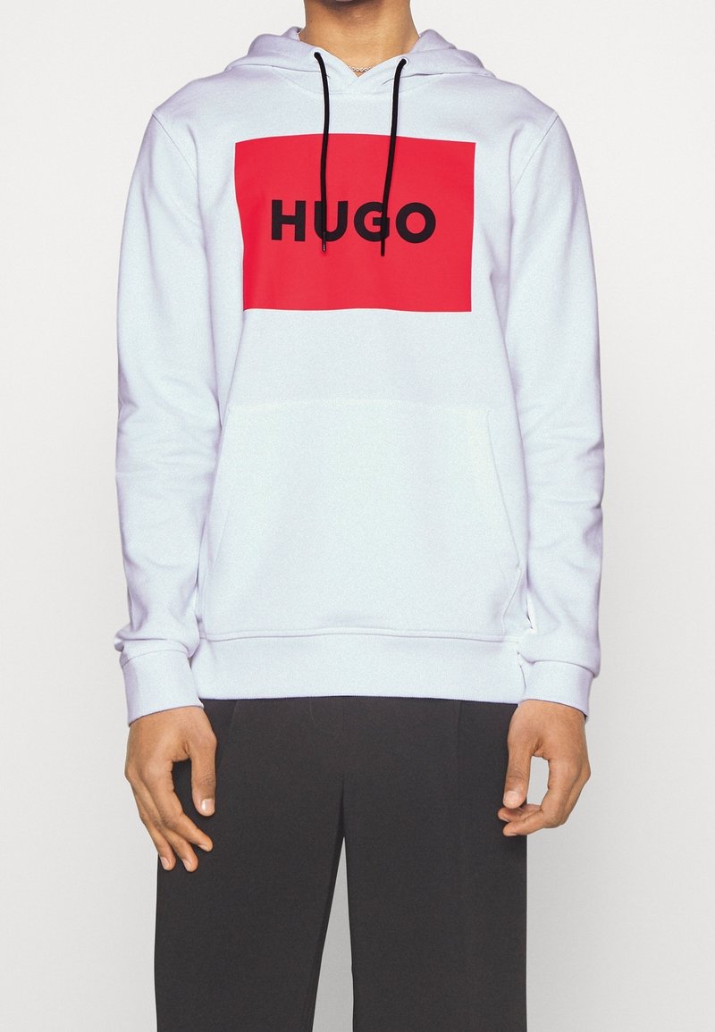 Sweat à capuche blanc avec un logo rectangulaire rouge comportant "HUGO" en lettres noires audacieuses. Cordons, poche kangourou et poignets côtelés.