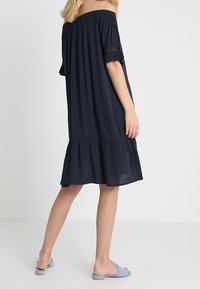 Marinblå avskuren klänning med korta volangärmar, en utsvängd nederkant och spetsdetaljer. I kombination med ljusblå sandaler.