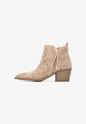 Botín beige de ante con patrones florales cortados con láser, cremallera dorada con borla, tacón de bloque de madera apilada y lengüeta en la parte trasera.