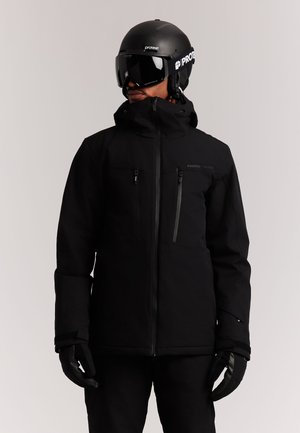 Personne portant une veste de ski noire, des gants, un casque et des lunettes, tournée légèrement vers la gauche sur un fond clair uni.